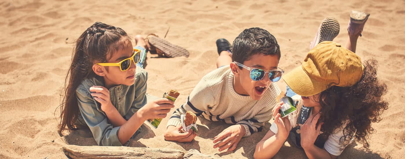 Three children lay in the sand eating Nature Valley bars and wearing sunglasses.
